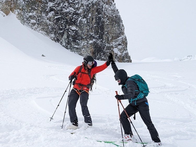 2 skiers high fiving each other with snow and rocky wall in background