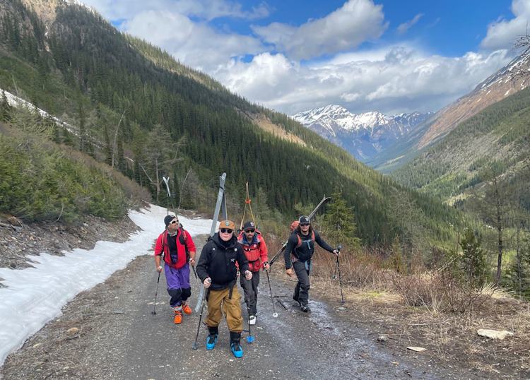 Four hikers walking on double track dirt road, all carrying skis on their backs and ski poles on their hands