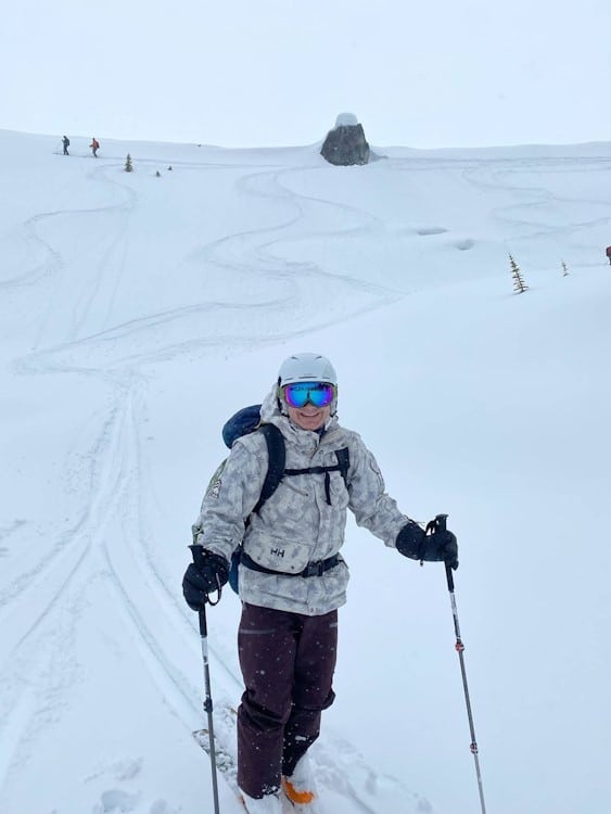 Skier smiling and posing in front of snowy hill with 2 skiers far away in the background