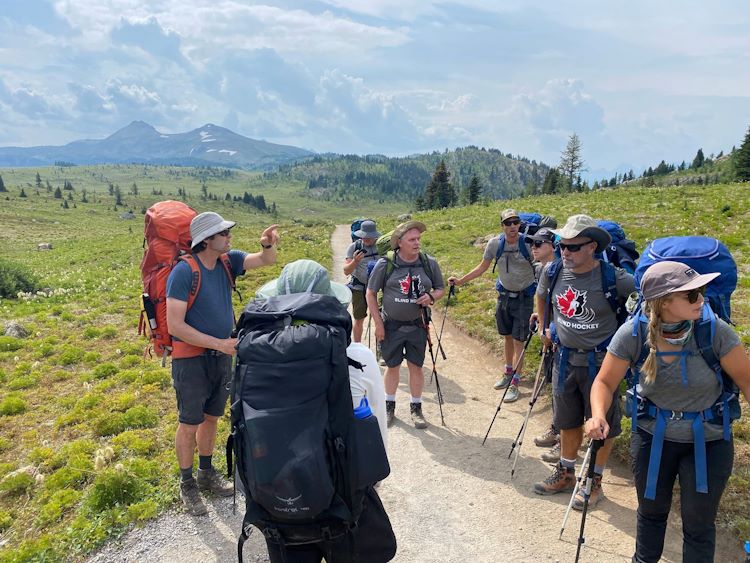 8 summer backpackers standing on trail with green valley and mountain in the background