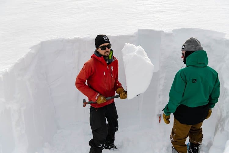 2 people standing in a hole in the snow with depth near their heads. One guy holding a shovel with tall piece of snow