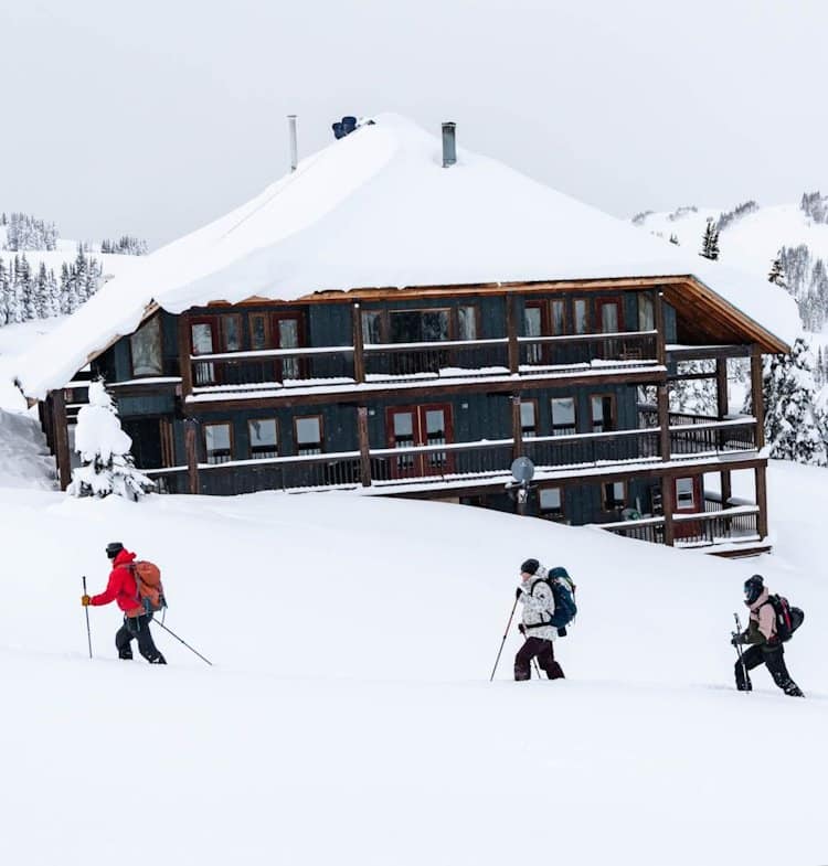 three skiers going across snow trail in front of a huge multi story hut in the winter