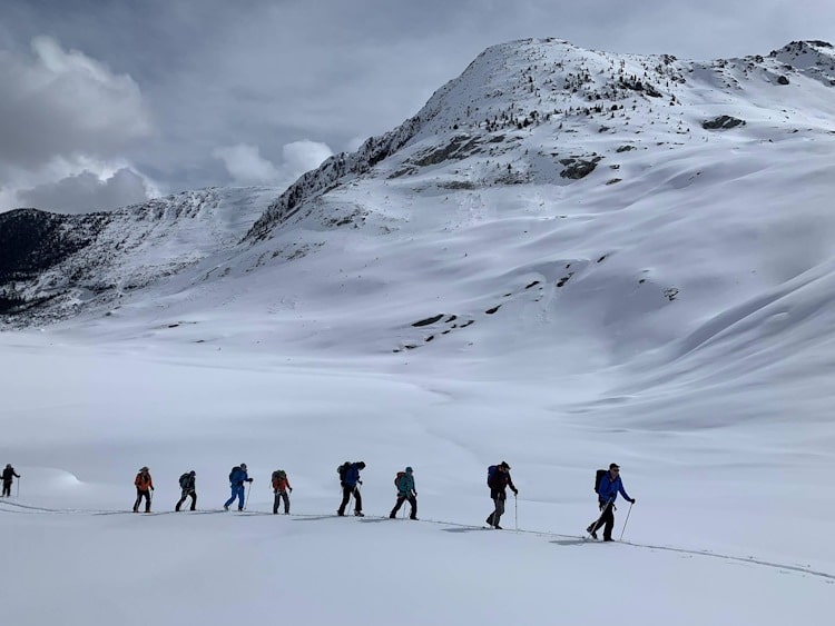 Nine backcountry skiers going along a flat snowy section with more snowy mountains in the background