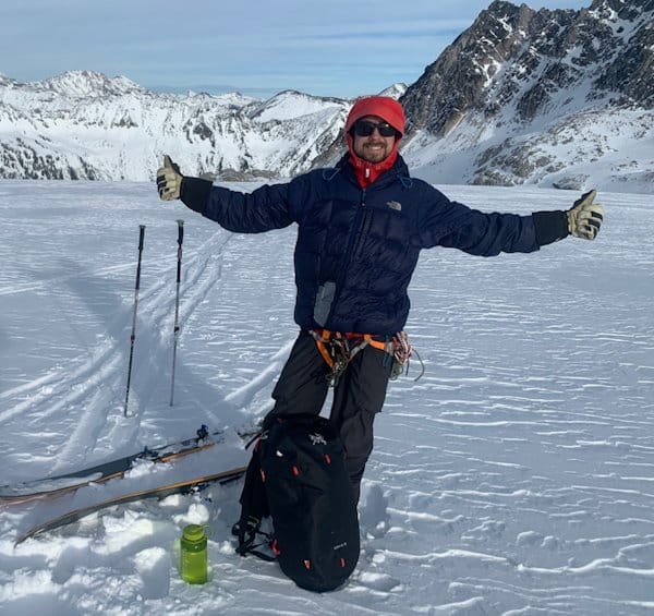 Man with two thumbs up pose standing in the a snowy mountainous backdrop.