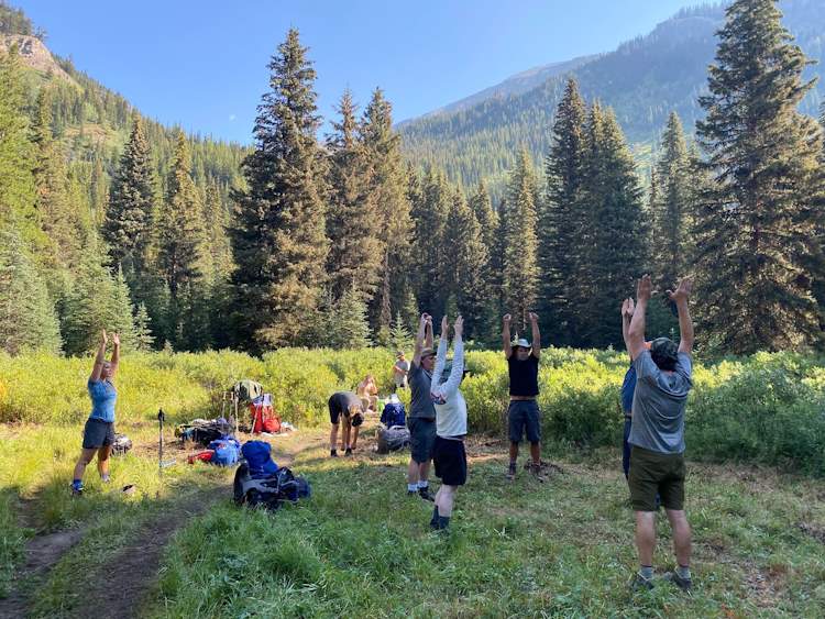 six hikers stretching hands up in the air with three other hikers milling around in a forest opening