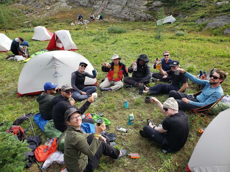 Ten people, including seven visually impaired individuals and three sighted guides, sit in a circle in a grassy field surrounded by one-person tents. The ten people each have a drink in their hand and are holding them up for "cheers". People's facial expressions look excited to be resting and enjoying down time after a full day of hiking up steep terrain.