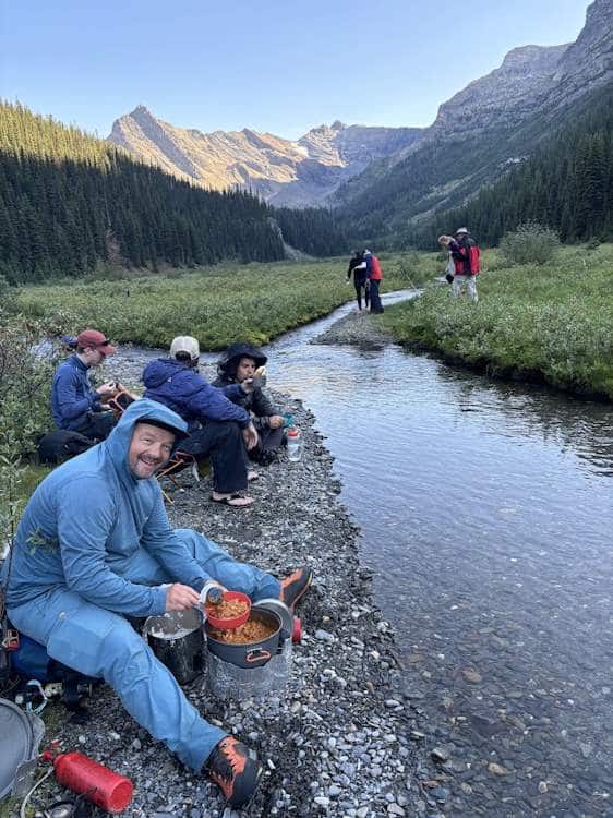 a group of people sitting at the edge of a small creek waiting for dinner to be prepared. The creek runs through an open meadow that's part grass and part ankle knee-high shrubbery.