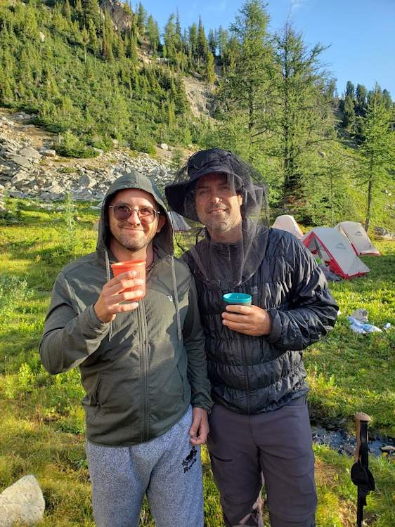 Two BMI participants, Jon and Mark, smile for a photo as they stand and enjoy their morning coffee out of their plastic camping mugs. Mark is wearing a brimmed bucket hat with a bug net overtop. Both are wearing warm clothes as the morning's sun just peaks over the nearby mountains.