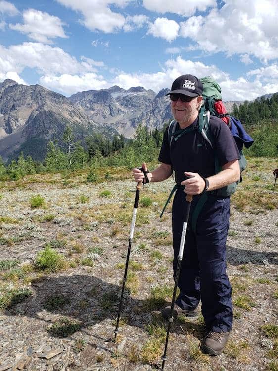 Blind participant, Ron who is in his early-60s, smiling for a photo on top of a ridge with the Septet Mountain Range visible in the background. A large glacier is off in the distance.