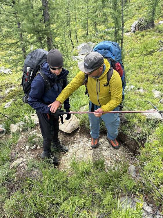 Playwest hiking guide Skinner, and visually impaired hiker, Jack taking a learning moment on the trail. The two are stopped and are checking out a bear dig that was likely created by a grizzly. Skinner is guiding Jack's hiking pole to help him locate the dig which is about 1 meter wide and half a meter deep.