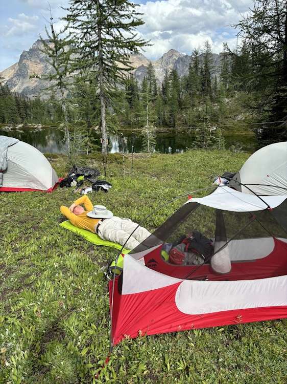 David, lying on his inflated sleeping pad on the grass next to his tent as he sets up his sleeping arrangement. David's body language implies that he is exhausted from the day of hiking. His arms are folded over his face to cover his eyes, and his brimmed sunhat is resting on his chest. In the background is Elsay Lake,