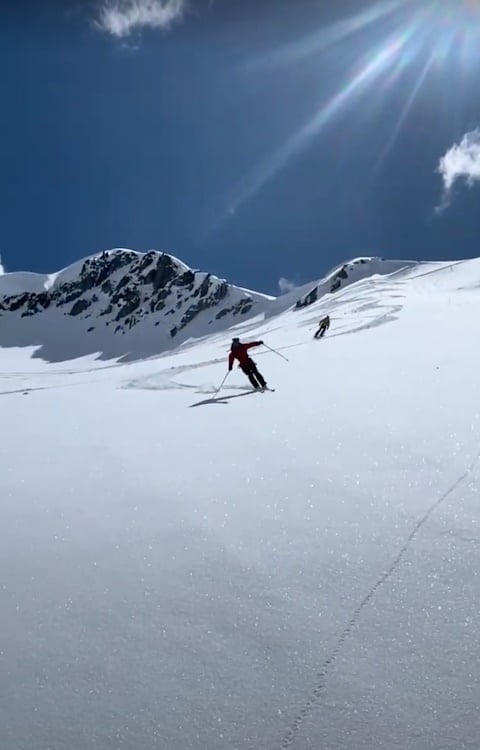 Two skiers with one main skier doing a casual turn on fresh steep powder with sun rays in the sky.