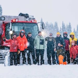 11 people in front of a snow cat vehicle in snowy outdoor environment with lots of trees in background.