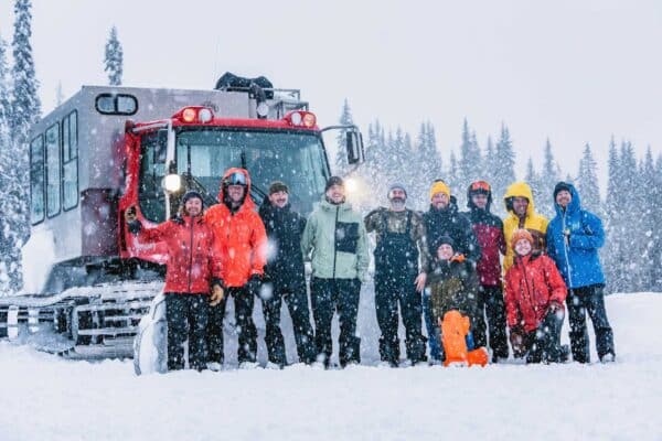 11 people in front of a snow cat vehicle in snowy outdoor environment with lots of trees in background.