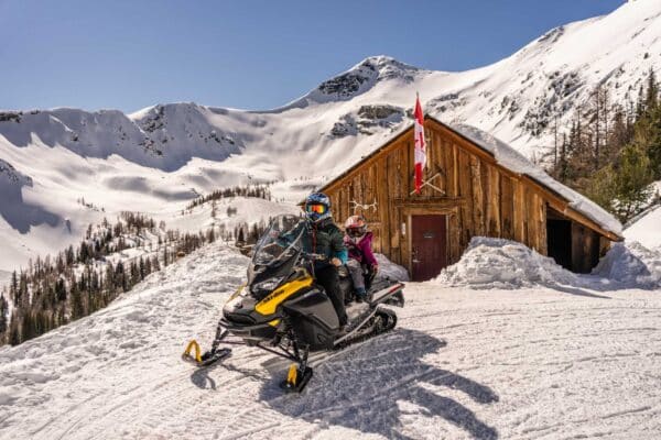 Clear sunny winter day mountainous scenery with wooden cabin and a snowmobile with helmeted adult and child.