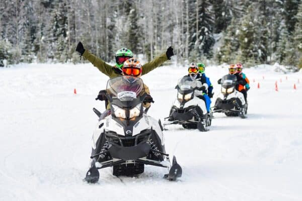 Couple of snowmobilers riding on one snowmobile with two other snowmobiles in the background with two riders each.