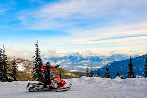 Snowmobiler standing on top of snowmobile on mountainous snowy environment.