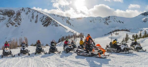 10 snowmobiles each with rider on a sunny winter day with mountains in the background.