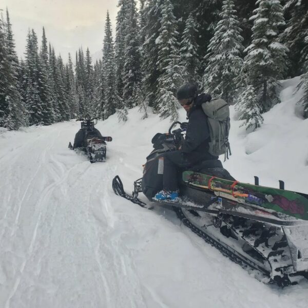 Two snowmobiles are seen heading up a snow-covered logging access road. The nearer sled has a Mad Max Sled Rack with a pair of skis attached.