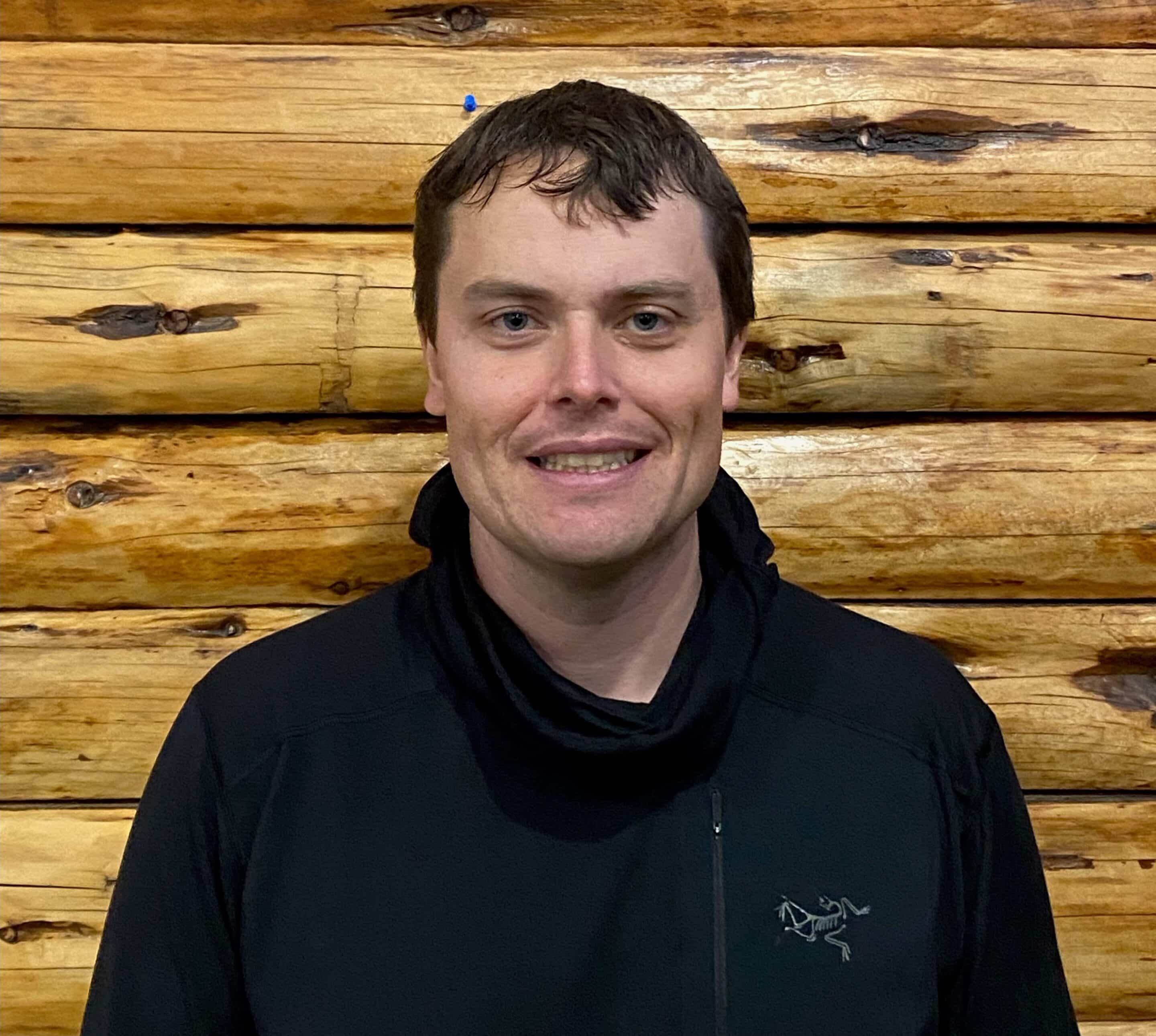 Headshot of Harry standing in front of a log-frame wall. Harry is in his early 30s, caucasian, and has short dark brown hair. He is wearing a black Arc'teryx jacket.