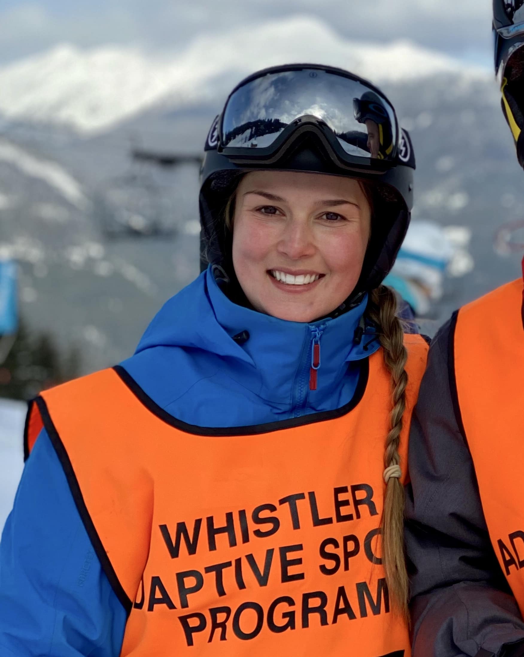 Alex smiles for a photo in her Whistler Adaptive Sports bright orange vest, overtop her ski gear. She is wearing a helmet and goggles, and is a caucasian in her early-30s.