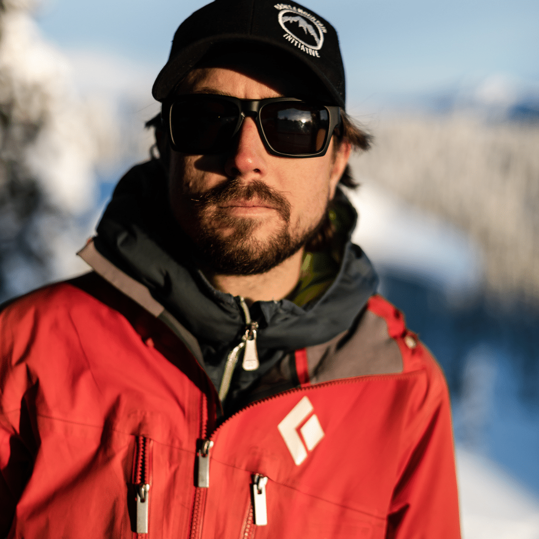 Tyson has a serious look on his face, wearing dark sunglasses, a BMI ball cap and a red Black Diamond shell jacket. Tyson has dark facial hair and is in his early-mid 30s.