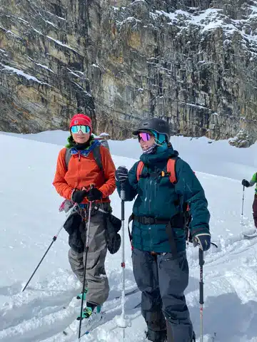 Sighted guide Alex stands in her ski gear next to a visually impaired skiier. They appear to be having a conversation, with a vertical rock wall in the background behind them.