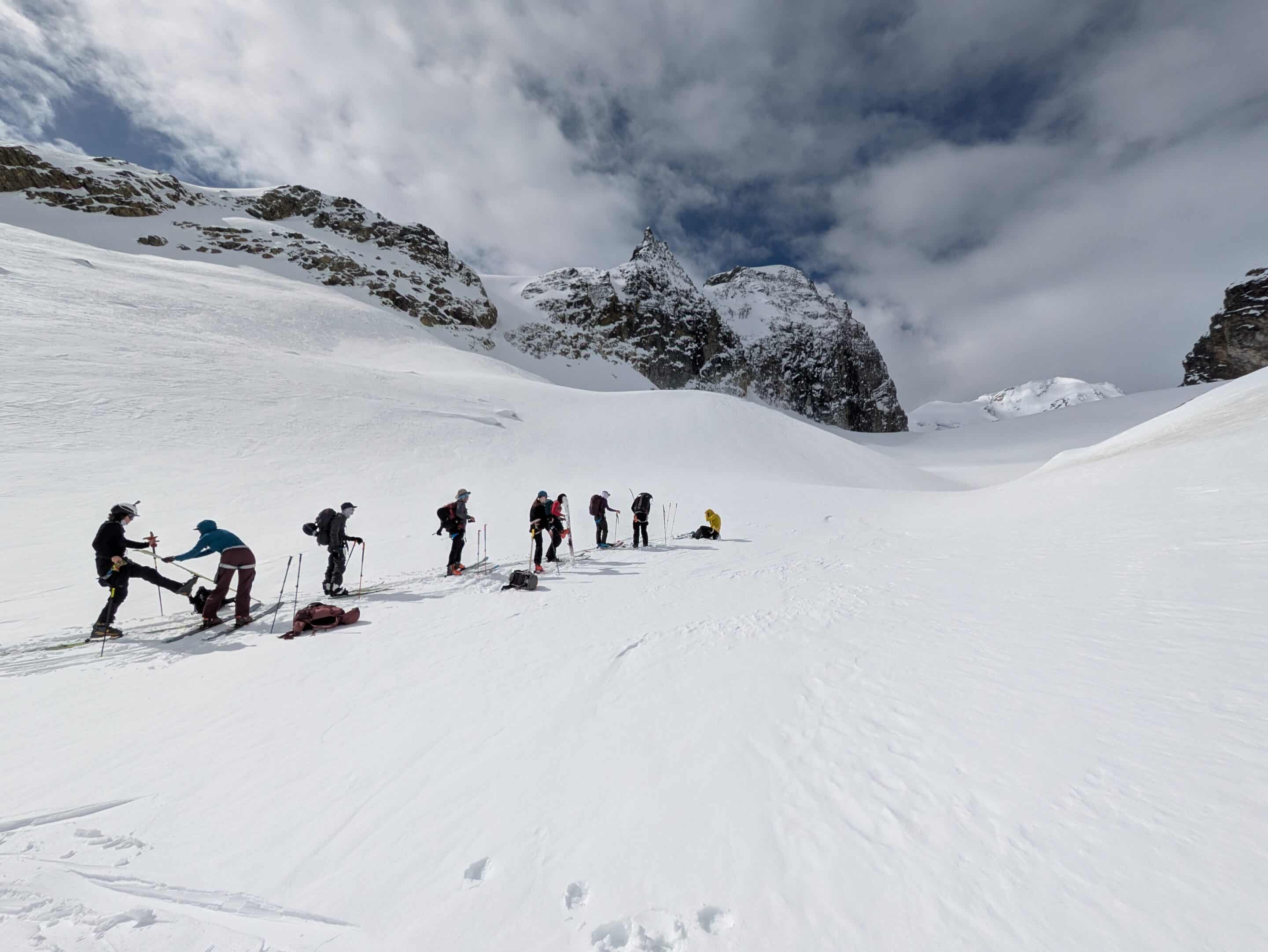 Seven people stand in a line on a skin track, spaced out for glacier travel, taking a quick break as they skin their way up Escargot Glacier.