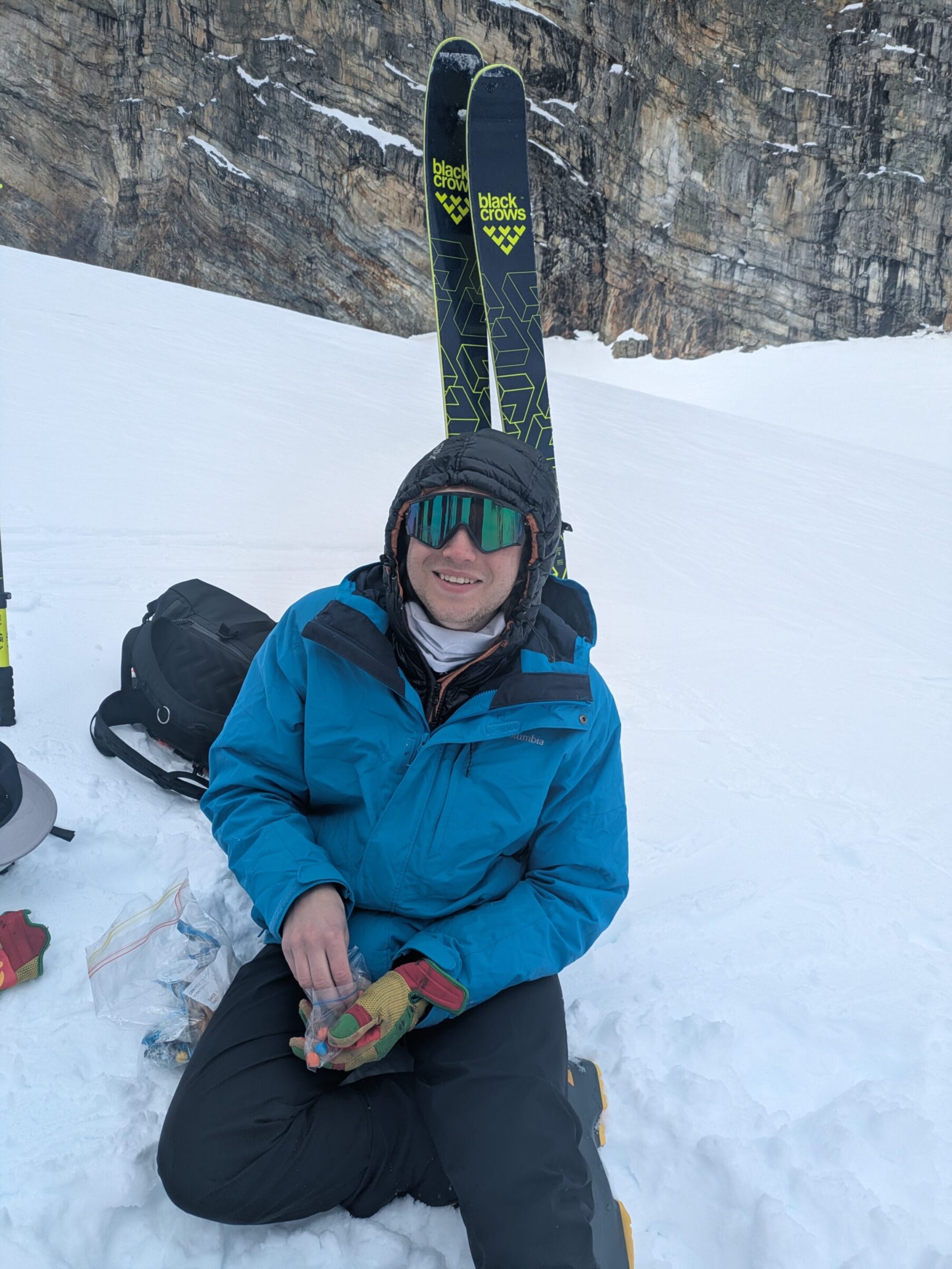 Ryan sits in the snow with his skis vertical in the snow providing a backrest, while he snacks on some trailmix.