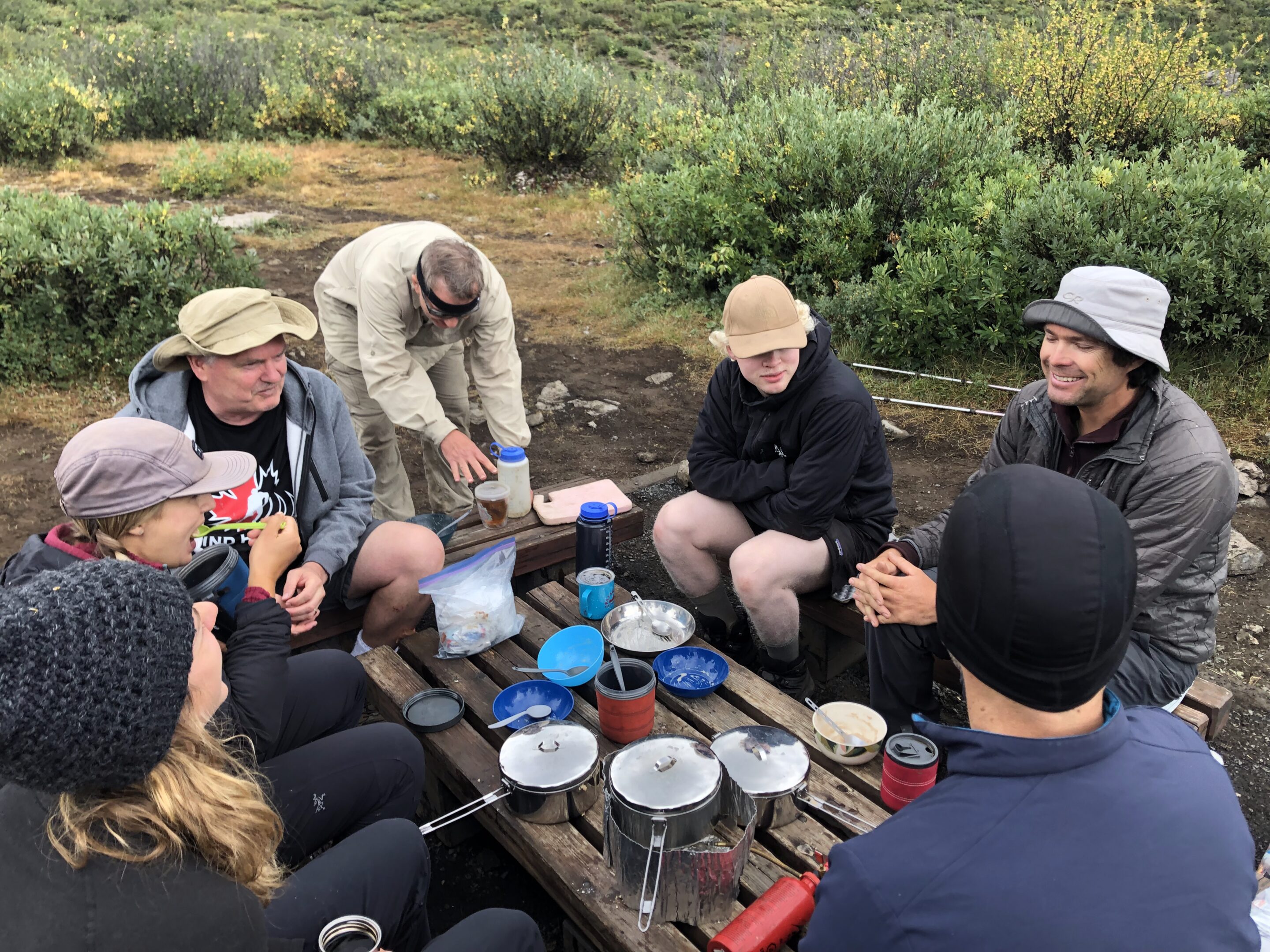 Seven people gather around a wooden picnic table sharing a meal.
