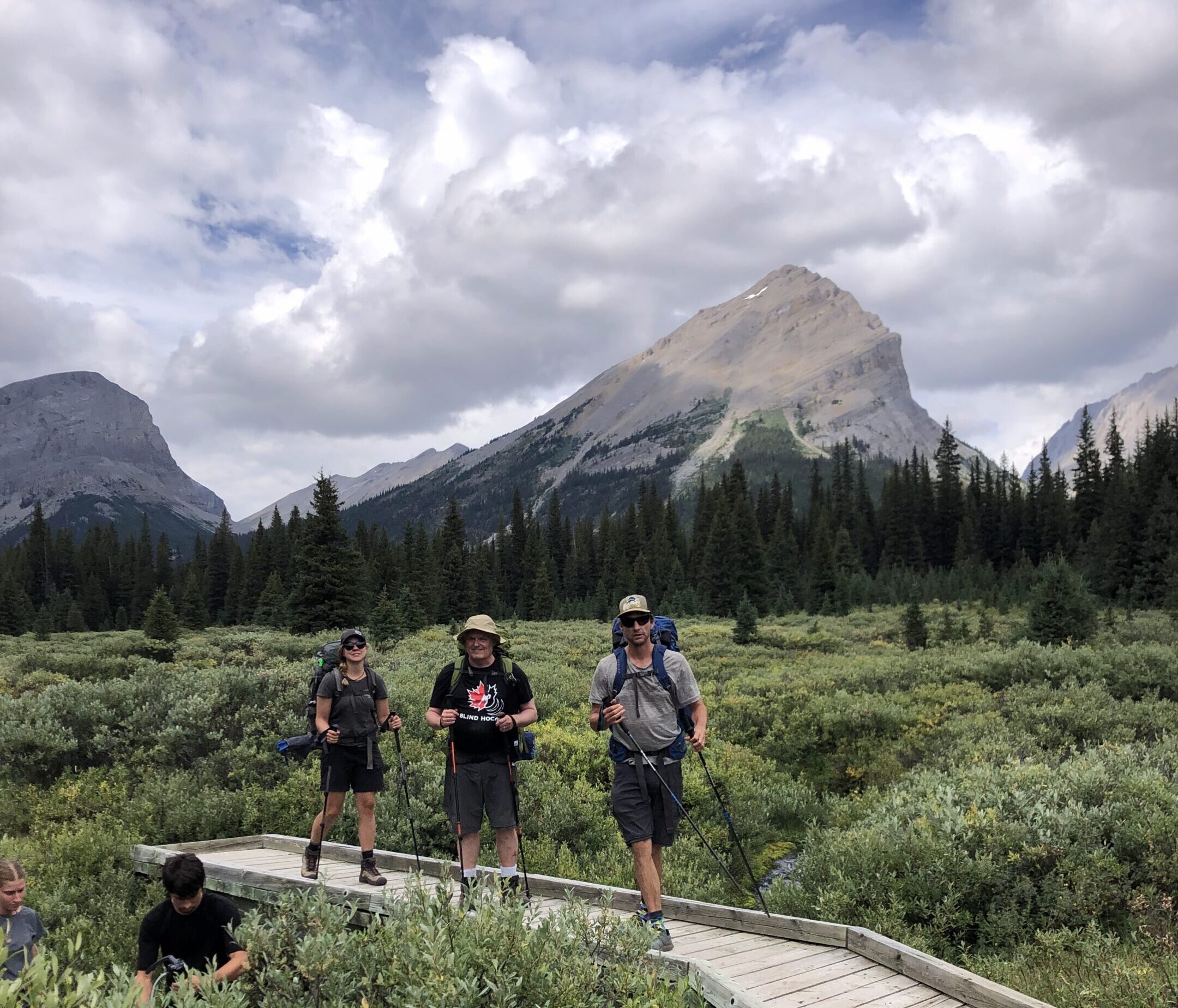 Three hikers cross a wooden bridge in a swampy meadow landscape, with a prominent mountain peak in the background.