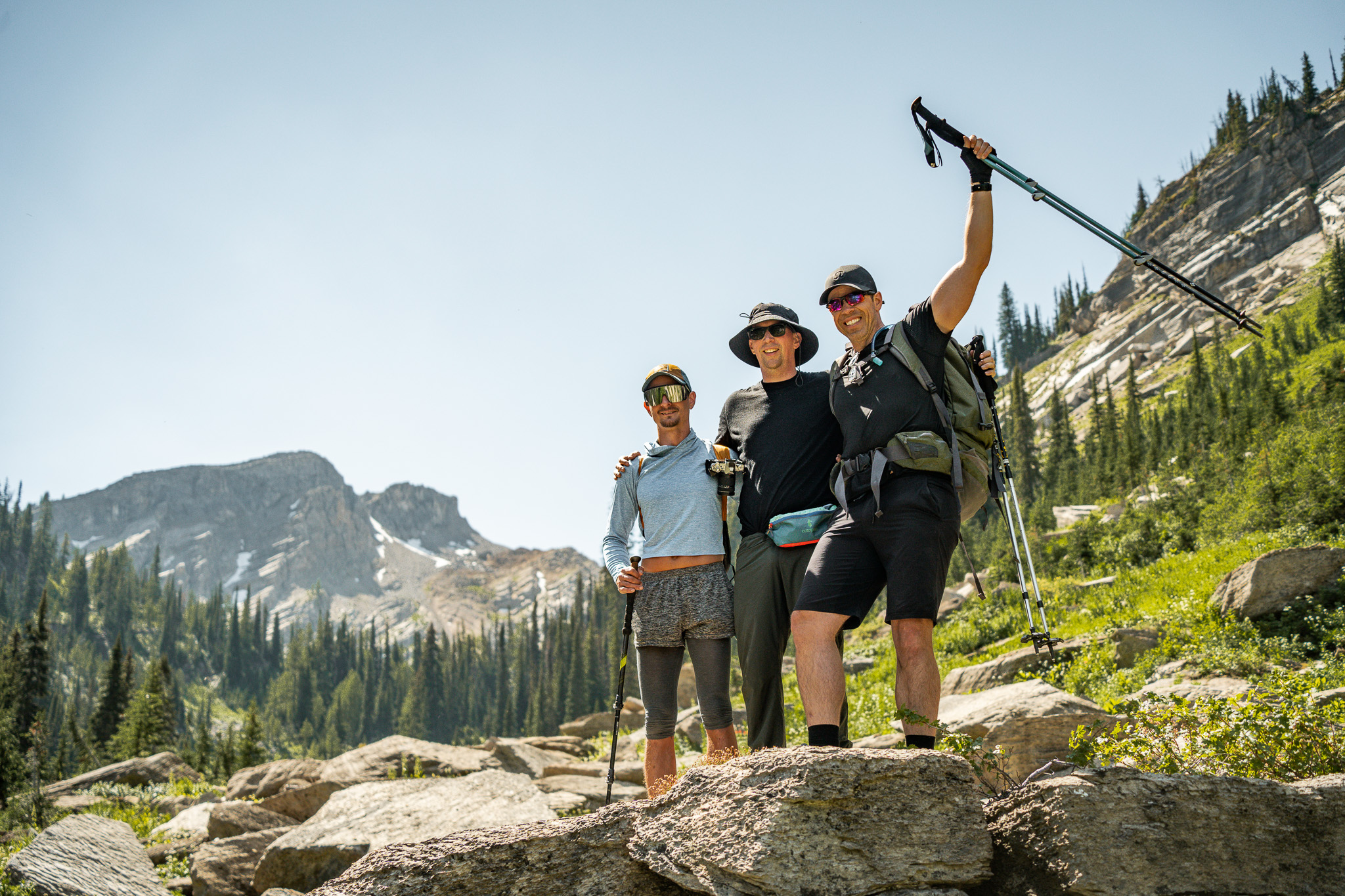 Stu (center), with sighted guide Josh (left), and fellow blind participant Lowell (right), pose holding trekking poles in the air, against a backdrop of evergreen forest and rugged mountain peaks - conveying a sense of accomplishment and camaraderie.