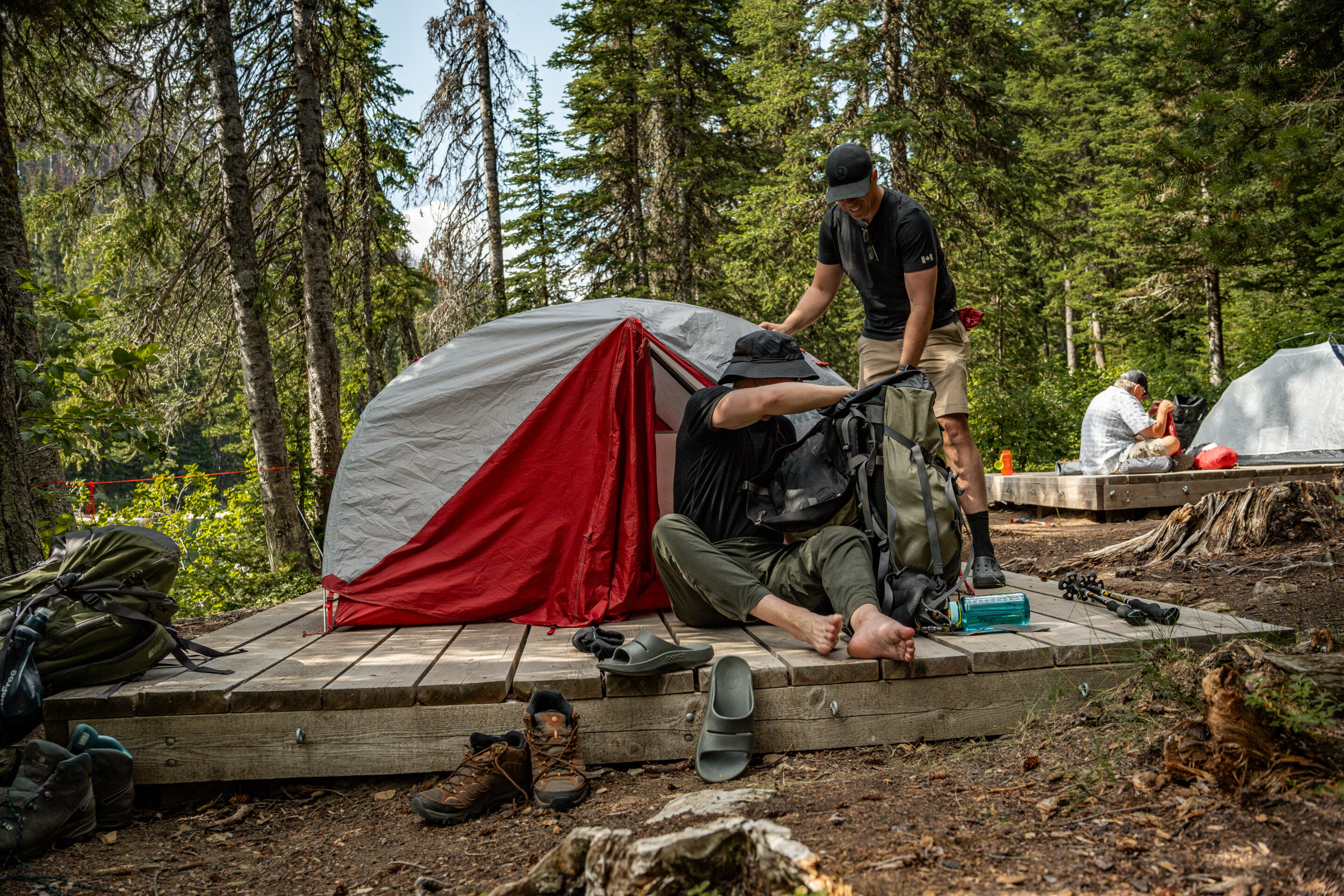 5 (1) Two people set up a tent at a forested backcountry campsite, while another person works at a nearby tent. Camping gear, backpacks, hiking boots, and trekking poles are scattered around the wooden tent platforms.