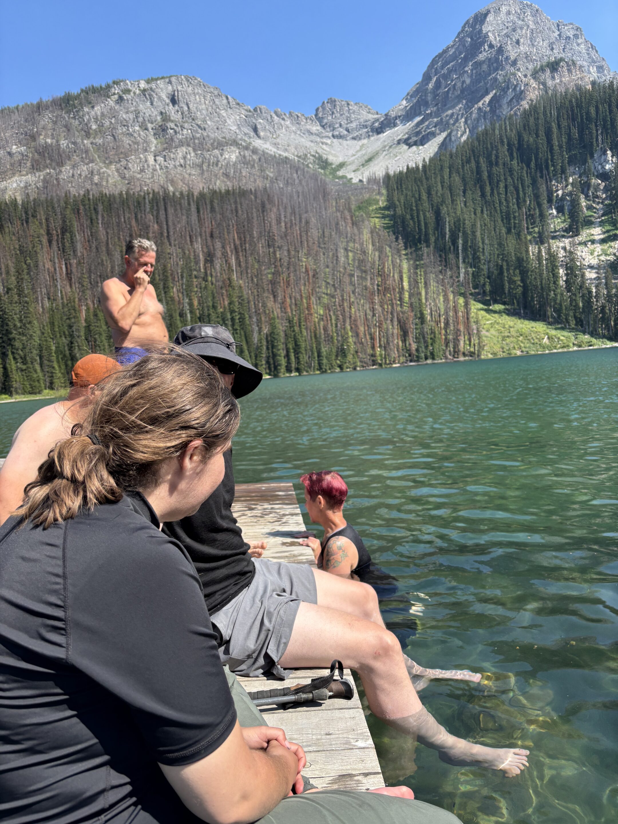 About seven individuals sit on a dock on a turquoise-coloured alpine lake, with one woman pictured swimming in the water.